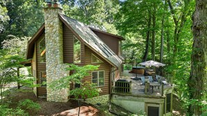 A log cabin with a stone chimney, a deck with outdoor furniture, and a forested backdrop. - Secluded Log Cabin Estate | 6 Private Acres | High-Altitude River Views (New Buffalo)
