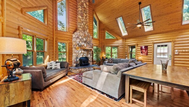 A log cabin interior with a stone fireplace, wooden furniture, and large windows. - Secluded Log Cabin Estate | 6 Private Acres | High-Altitude River Views (New Buffalo)