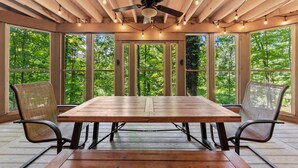A wooden dining table with chairs, large windows, and a ceiling fan in a room with a view of trees. - Secluded Log Cabin Estate | 6 Private Acres | High-Altitude River Views (New Buffalo)