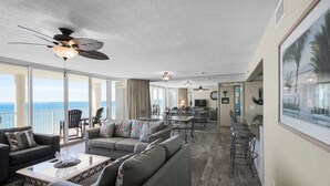 A spacious living area with a large sofa, a coffee table, and a dining table set for four. A ceiling fan and a large window offering a view of the ocean are visible. - Oceanfront 2BR condo in Panama City Beach, sleeps up to 8, near attractions. (Panama City Beach)