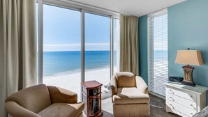 A hotel room with a large window offering a beach view, two armchairs, a wooden cabinet, and a bedside table with a lamp. - Oceanfront 2BR condo in Panama City Beach, sleeps up to 8, near attractions. (Panama City Beach)