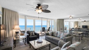 A spacious living room with a large sofa, a coffee table, and a dining area with chairs and a table. There is a ceiling fan and a TV mounted on the wall. - Oceanfront 2BR condo in Panama City Beach, sleeps up to 8, near attractions. (Panama City Beach)