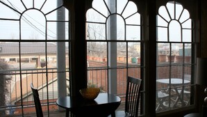 Ein Zimmer mit Tisch und Stühlen neben großen Bogenfenstern, die auf einen Innenhof mit einem Backsteingebäude und einer grünen Mülltonne blicken.