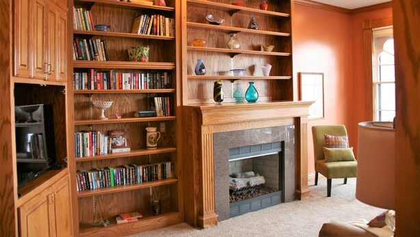 Eine Bibliothek mit mehreren Holzbücherregalen, gefüllt mit Büchern und dekorativen Gegenständen, einem Kamin mit brennendem Holz und einem grünen Sessel.