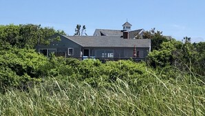 Een groot huis met een opvallende toren, omgeven door weelderige begroeiing en hoog gras.