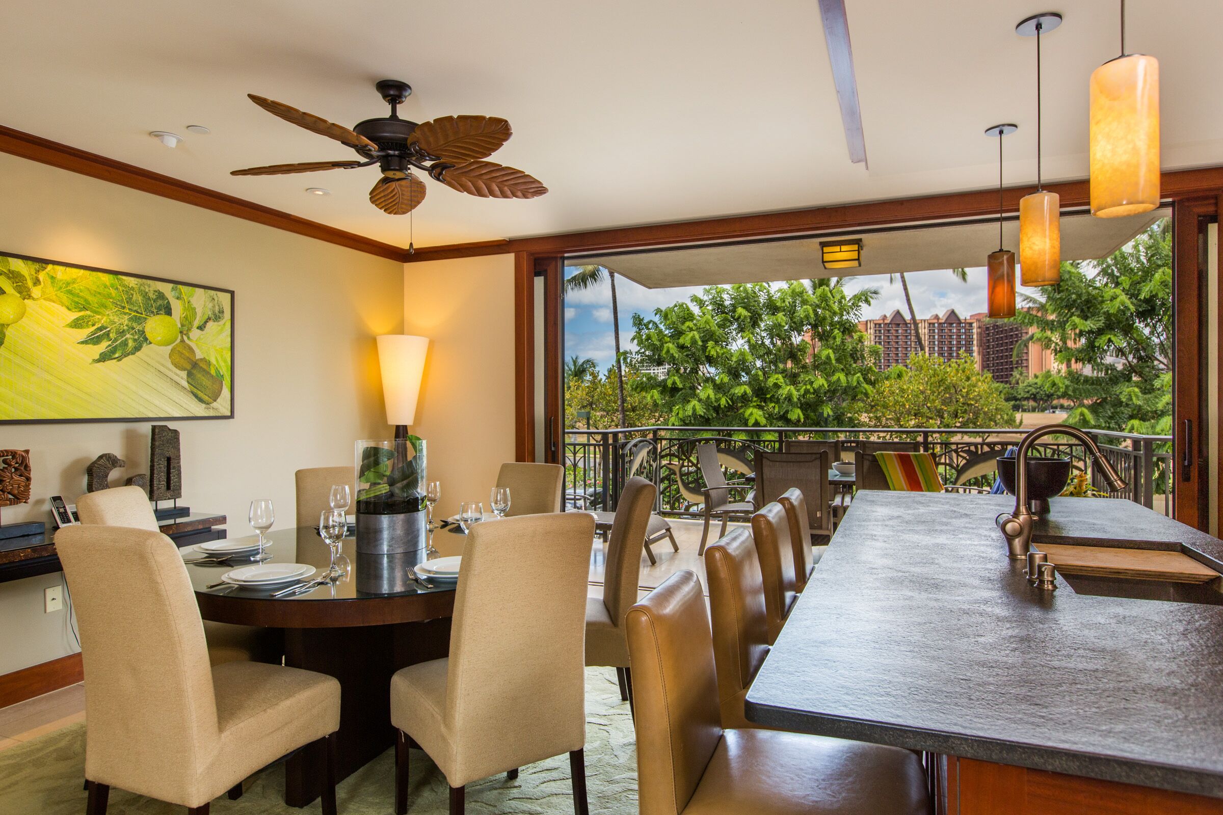 A dining area with a large table, chairs, a ceiling fan, and a view of greenery and buildings.