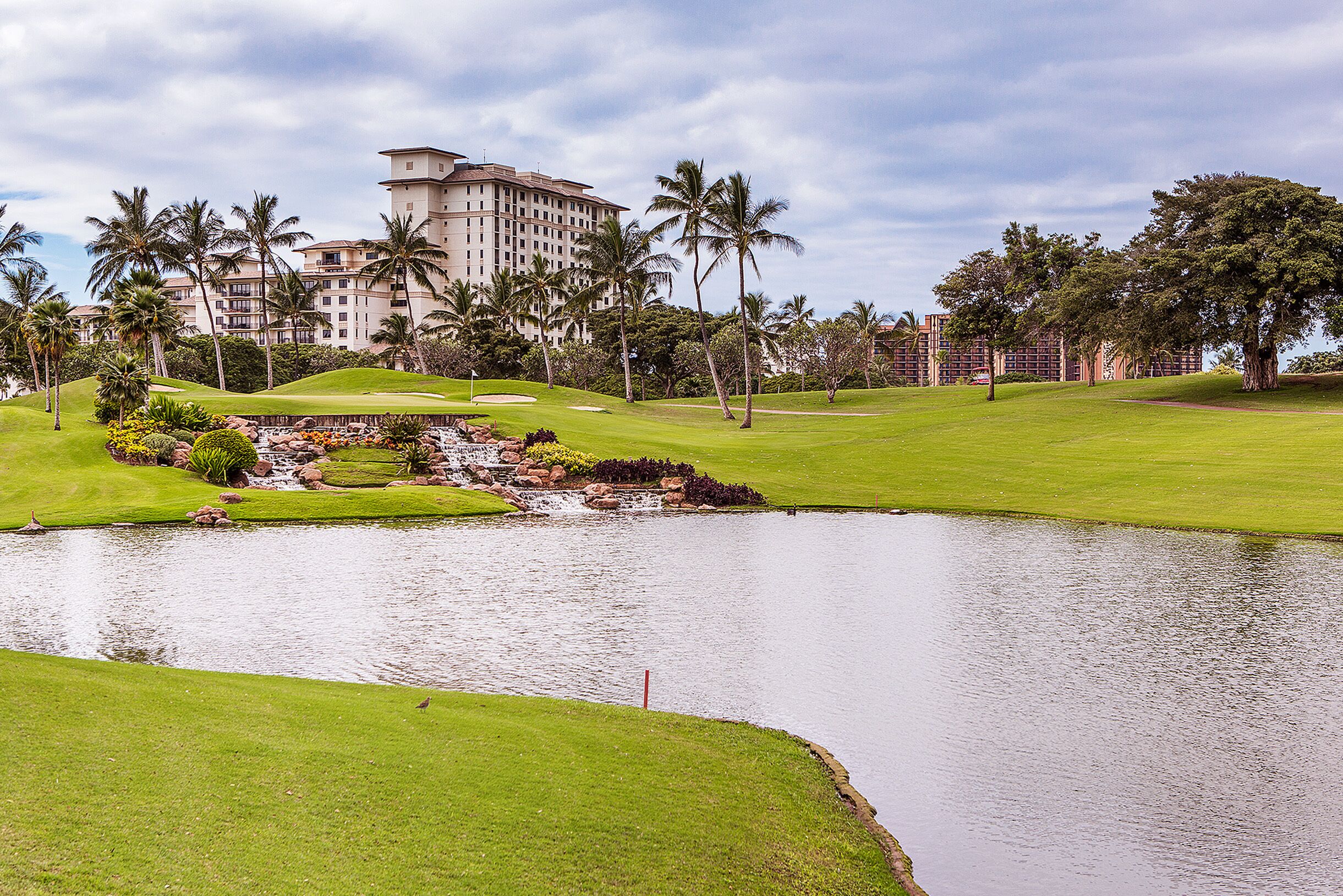 Un campo de golf con un estanque, palmeras y un hotel grande al fondo.