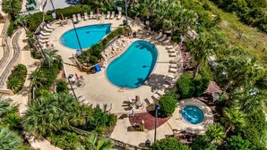 An aerial view of a resort with two large swimming pools surrounded by lounge areas and palm trees.