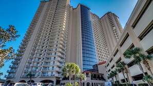 A high-rise hotel with multiple balconies, a parking lot, and palm trees.