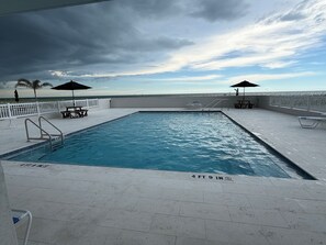 Une piscine rectangulaire dotée d’une terrasse blanche, entourée de rampes et de parasols blancs, et offrant une vue sur la mer et un ciel nuageux.