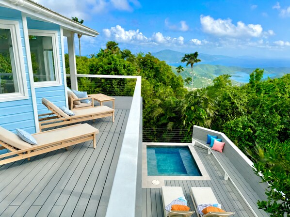 A deck with lounge chairs and a hot tub overlooking a lush landscape and a body of water.