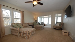 A living room with a sectional sofa, ottoman, and armchair, large windows, ceiling fan, and a flat-screen TV mounted on the wall. - Lake House with Sandy Beach and Amazing Sunsets (Delton)