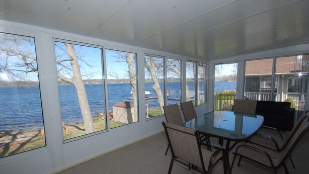 A dining area with a glass table and chairs, large windows offering a view of a lake and trees. - Lake House with Sandy Beach and Amazing Sunsets (Delton)