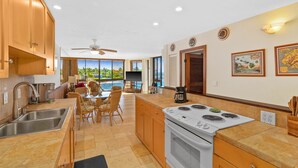 A modern kitchen with a large island, stainless steel sink, and a white stove. There is a dining area with a table and chairs, and a view of the ocean through large windows. - Spectacular Oceanfront Corner Unit Condo at Kuhio Shores (Koloa)