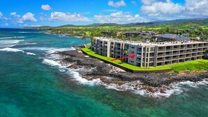 A hotel with multiple floors, located on a rocky coastline with clear turquoise waters. - Spectacular Oceanfront Corner Unit Condo at Kuhio Shores (Koloa)