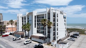 A multi-story hotel with a white facade, located near a beach with palm trees and a parking lot. - Seas Life's Moments - Condo 202: Spacious 2BR retreat in South Padre Island, sleeps 7. (South Padre Island)
