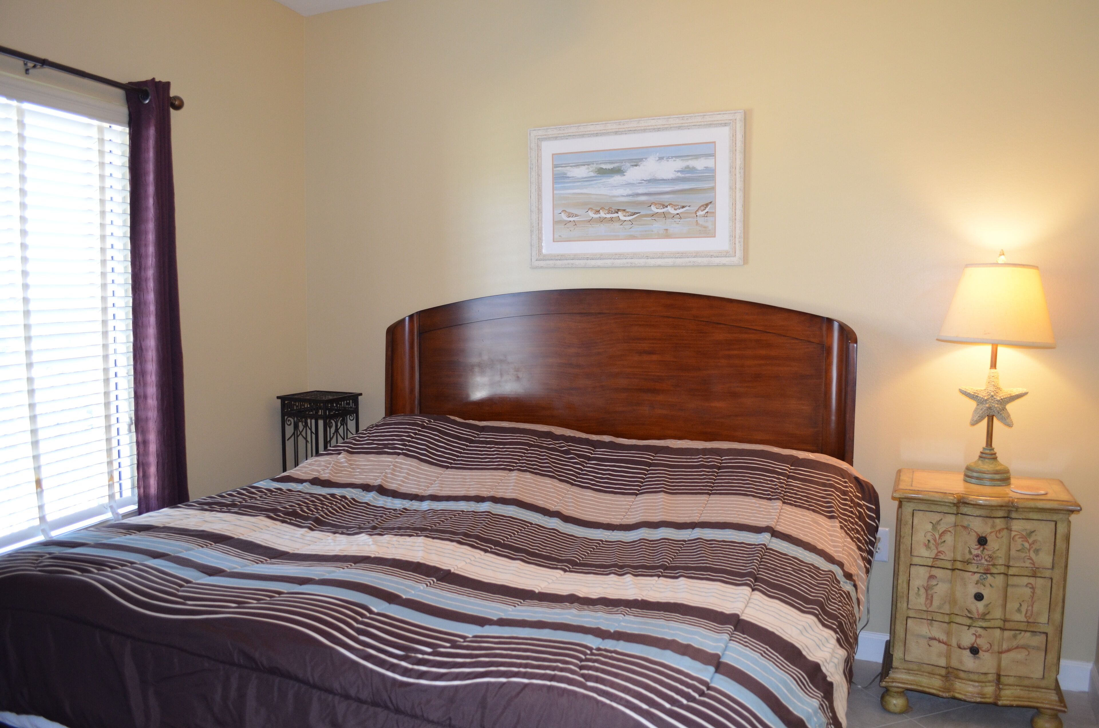 A bedroom with a wooden headboard, a striped bedspread, a nightstand with a lamp, and a framed picture on the wall.