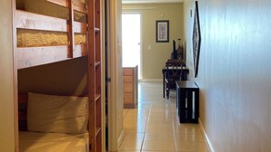 Un pasillo con una cama litera a la izquierda, una zona de comedor con sillas y una mesa en el centro, y una puerta que da a otra habitación al final.