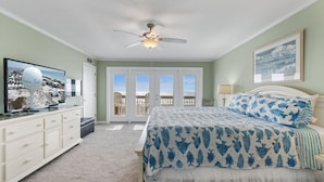 A bedroom with a bed, a dresser, a ceiling fan, a television, and a view of the beach through the sliding glass doors.