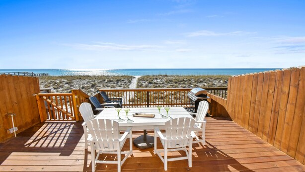 A wooden deck with a table and chairs, overlooking a beach with a clear view of the ocean.