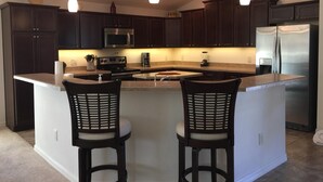 A modern kitchen with dark wood cabinets, stainless steel appliances, a central island with two bar stools, pendant lighting, and a clock on the wall. - 2 BED/2BATH PLUS DEN  COURTYARD VILLA (The Villages)