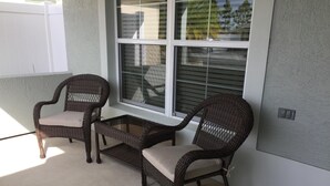 A patio area with two wicker chairs and a small table, a white window with blinds, and a concrete floor. - 2 BED/2BATH PLUS DEN  COURTYARD VILLA (The Villages)