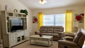 A living room with a flat-screen TV on a white cabinet, a beige sofa, a coffee table, and yellow curtains. - 2 BED/2BATH PLUS DEN  COURTYARD VILLA (The Villages)