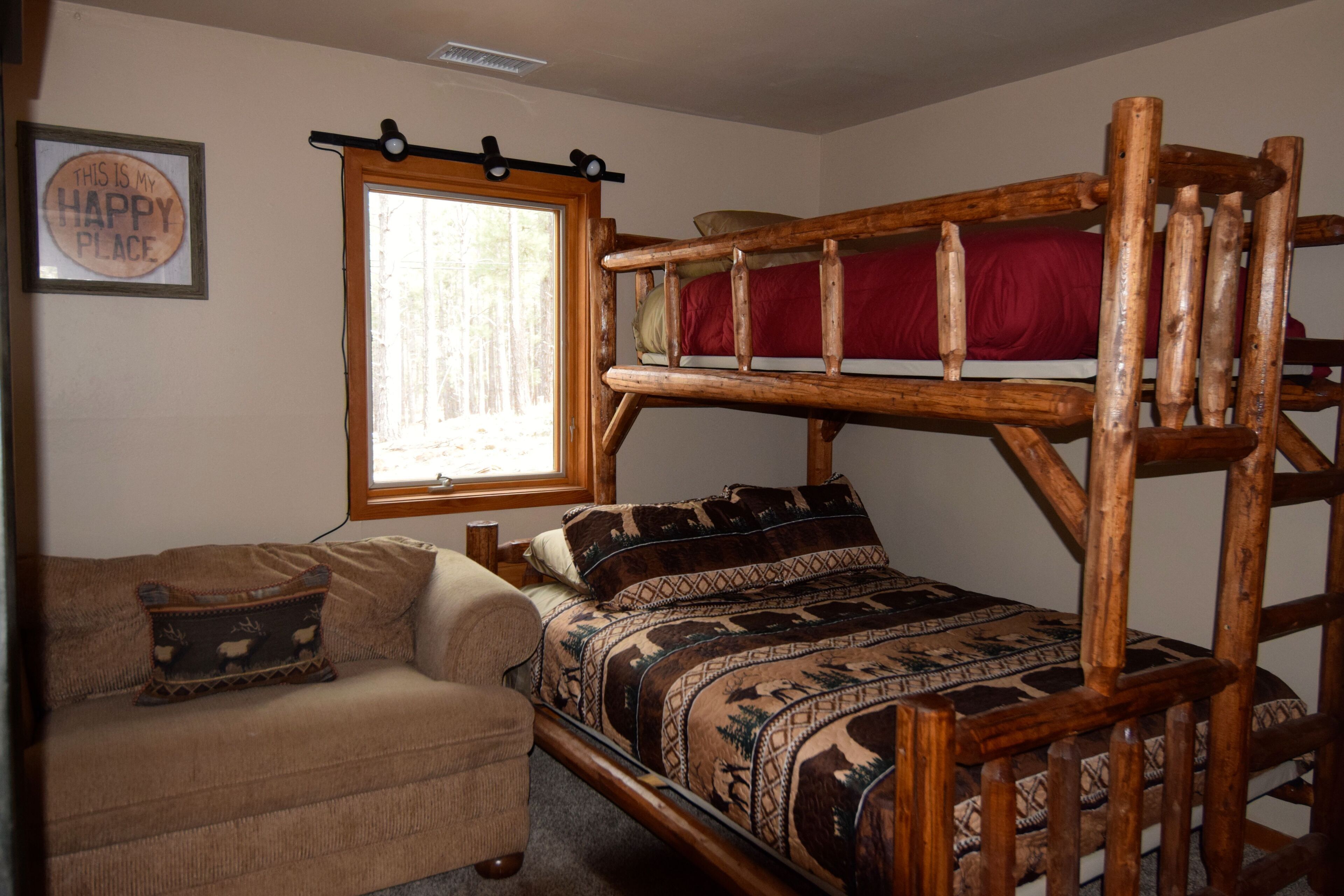 Un lit superposé avec un matelas rouge, une tête de lit en bois et une couverture à motifs. Il y a une fenêtre donnant sur des arbres, un canapé avec un coussin à motifs et un panneau accroché au mur.