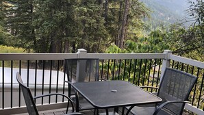 Een terras met een tafel en stoelen, omgeven door een witte reling, met uitzicht op een bebost gebied en bergen in de verte.