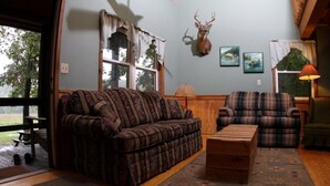A cozy living room with a patterned sofa, a plaid armchair, and a wooden coffee table. A deer head mounted on the wall adds a rustic touch. - Getaway Cabin next door to Maramec Spring Park & Meramec River Trophy Trout (St James)