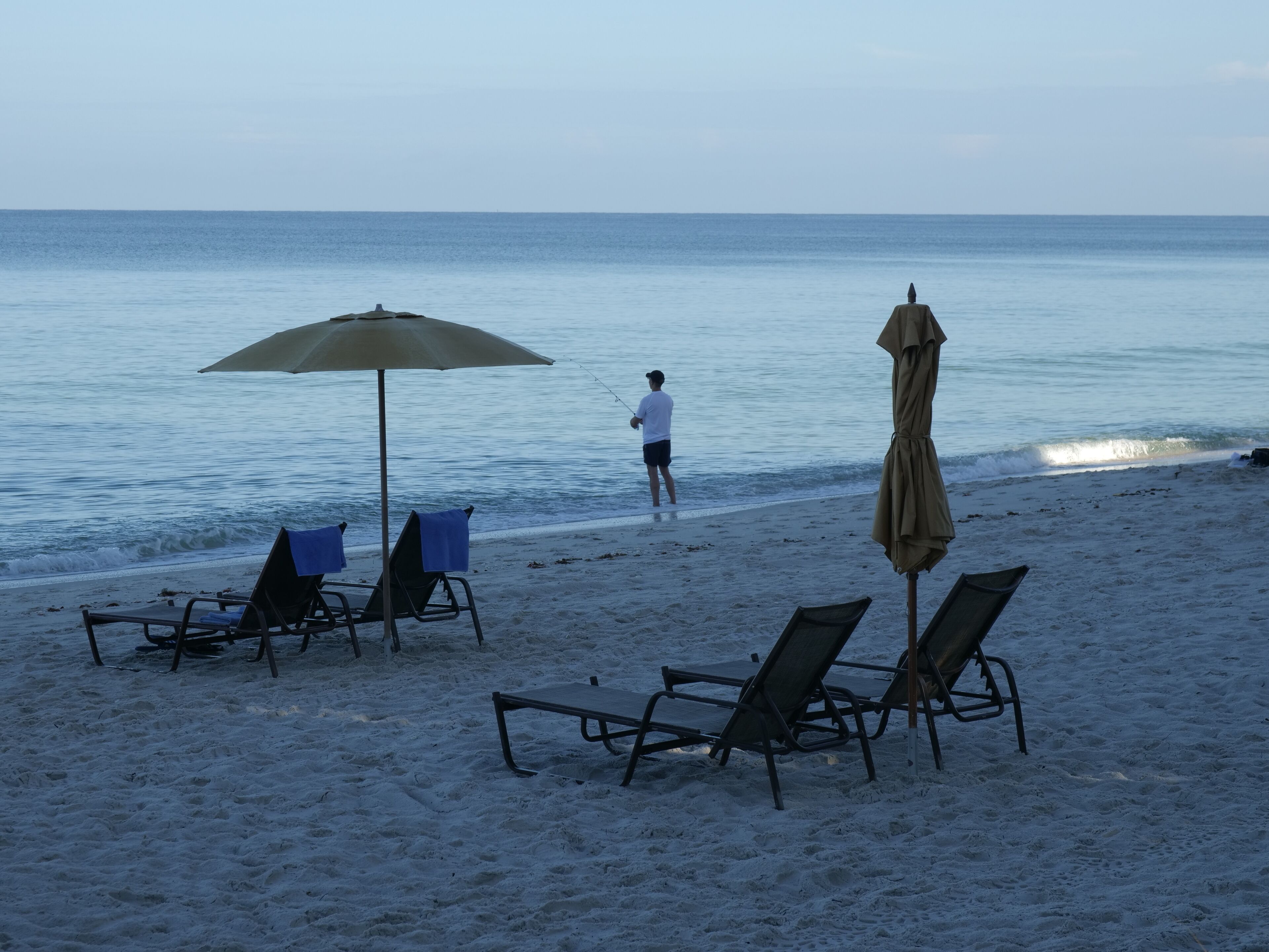 A beach with lounge chairs and umbrellas, a person fishing, and calm sea waves.