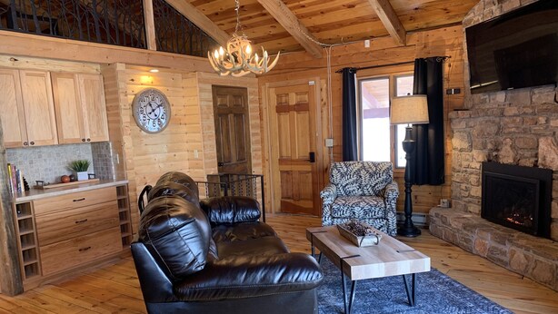 Un acogedor interior de cabaña de troncos con un sofá de cuero, un sillón estampado, una mesa de centro de madera, una chimenea de piedra y un reloj grande en la pared.