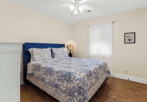 Un dormitorio con cabecera azul, colcha estampada, ventilador de techo, lámpara de noche y ventana con persianas.