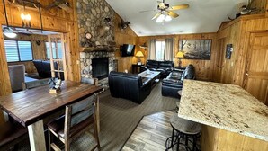 Ein rustikales Ferienhäuschen mit Kamin aus Stein, Holzwänden, einem Essbereich, einem Wohnzimmer mit Sofa und Fernseher sowie einer Küche mit Granitarbeitsplatten.