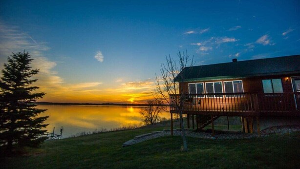 Eine Blockhütte am Seeufer mit Terrasse, Blick auf den See und einem Sonnenuntergang.