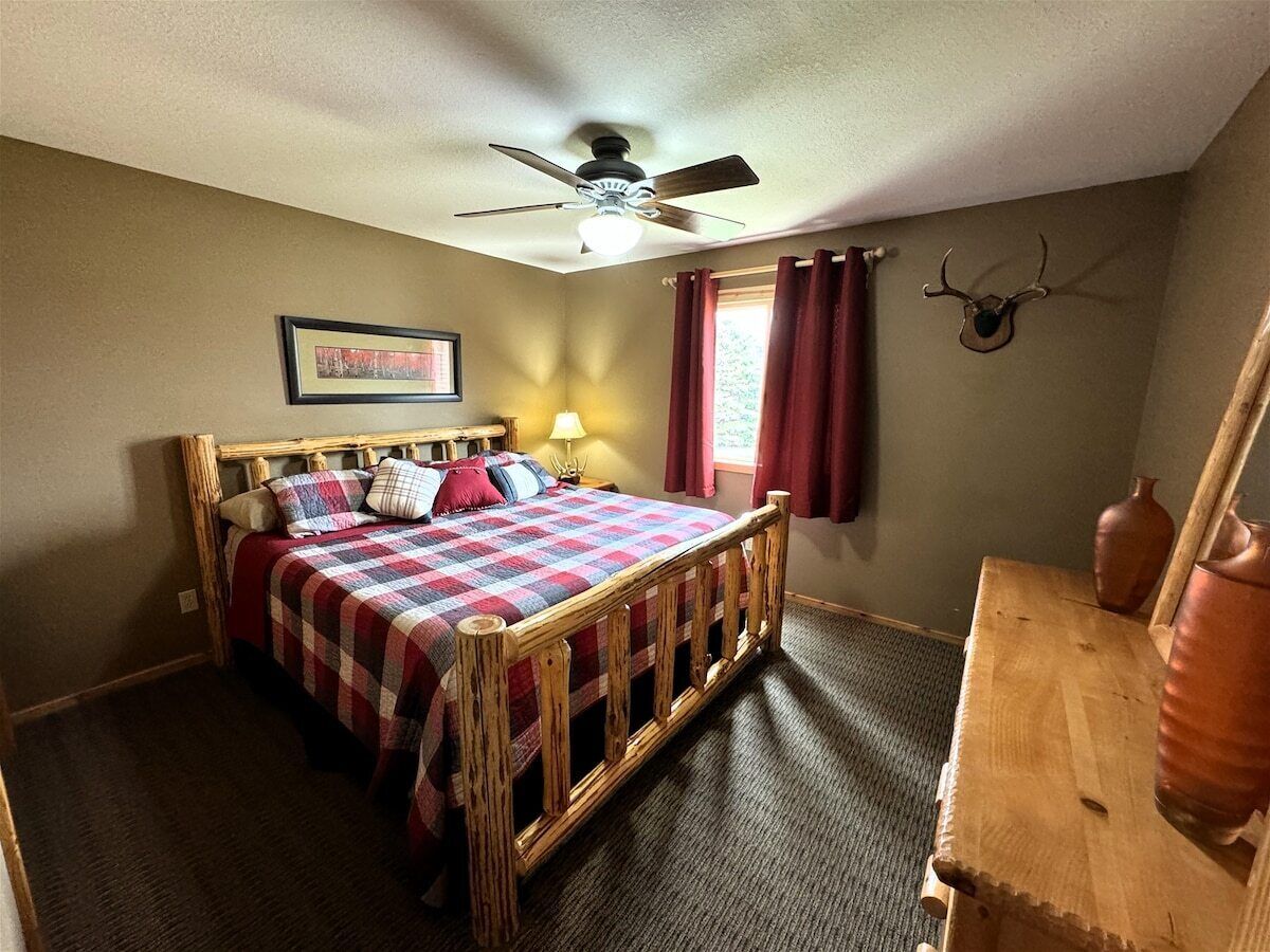 A bedroom with a wooden bed, a ceiling fan, a window with red curtains, and a wall-mounted deer head.