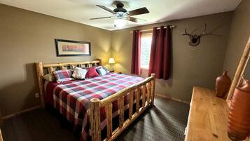 A bedroom with a wooden bed, a ceiling fan, a window with red curtains, and a wall-mounted deer head.