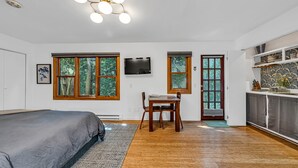 A bedroom with a bed, a desk, and a TV. There are windows and a door leading to the kitchen.