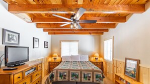 Une chambre avec un plafond en bois, un lit recouvert d’une courtepointe, des lampes de chevet, une télévision, une bibliothèque et des tableaux encadrés accrochés au mur.