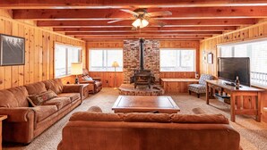 Un intérieur de chalet chaleureux avec des murs en bois, une cheminée, un ventilateur de plafond et un grand téléviseur.