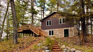A cabin with a stone exterior, wooden deck, and a staircase leading to the entrance. - Lakeside Cottage in the Pines with Huge Deck, 40-foot Dock, and Sunset Views (Hazelhurst)