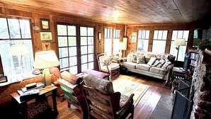 A cozy cabin interior with wooden walls, a sofa, a dining table, and a fireplace. - Lakeside Cottage in the Pines with Huge Deck, 40-foot Dock, and Sunset Views (Hazelhurst)
