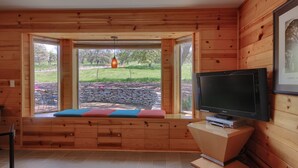 Interior d'una cabana de fusta amb un gran finestral, una televisió i un banc amb coixins.