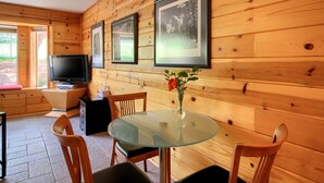 Interior d'una cabana de fusta amb una taula de vidre, cadires de fusta, un televisor i quadres amb marc a la paret.