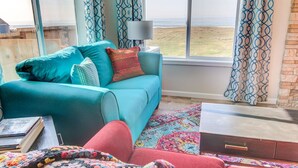 A room with a teal sofa, a patterned rug, a wooden side table with a lamp, and a view of the beach.