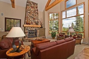 A cozy living room with a stone fireplace, leather sofas, and large windows offering a view of trees.
