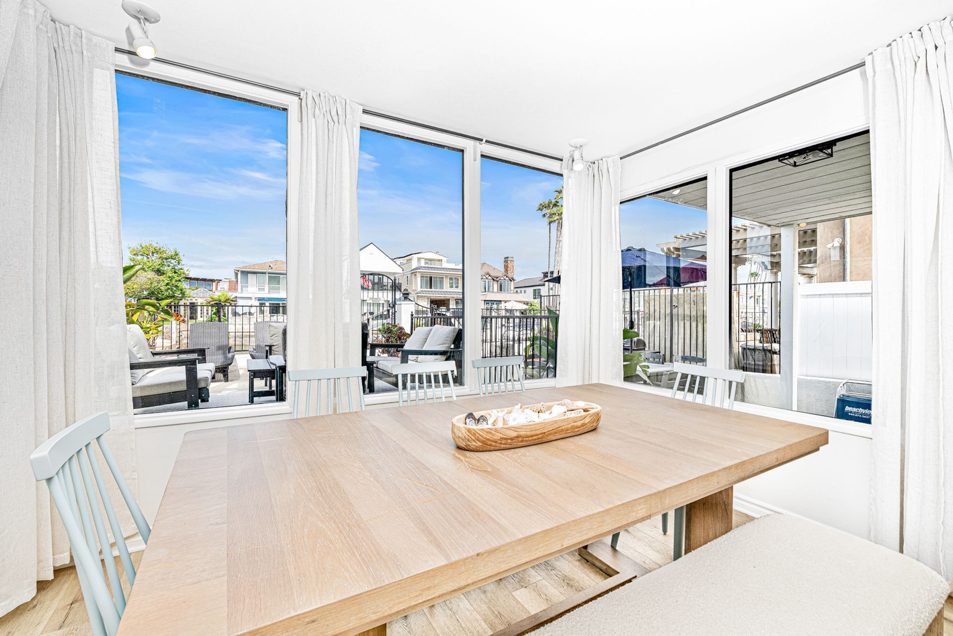 A spacious balcony with a wooden table, white curtains, and large windows offering a view of neighboring houses.