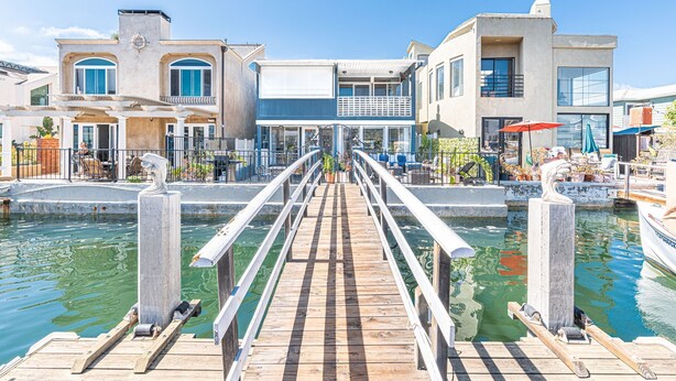A wooden dock leading to a waterfront property with a pool, a covered patio, and a view of the water.