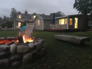 A house with a stone fire pit, a deck with outdoor furniture, and a view of the surrounding neighborhood. - The Narrows Yacht Club Cottage (Lake Leelanau)