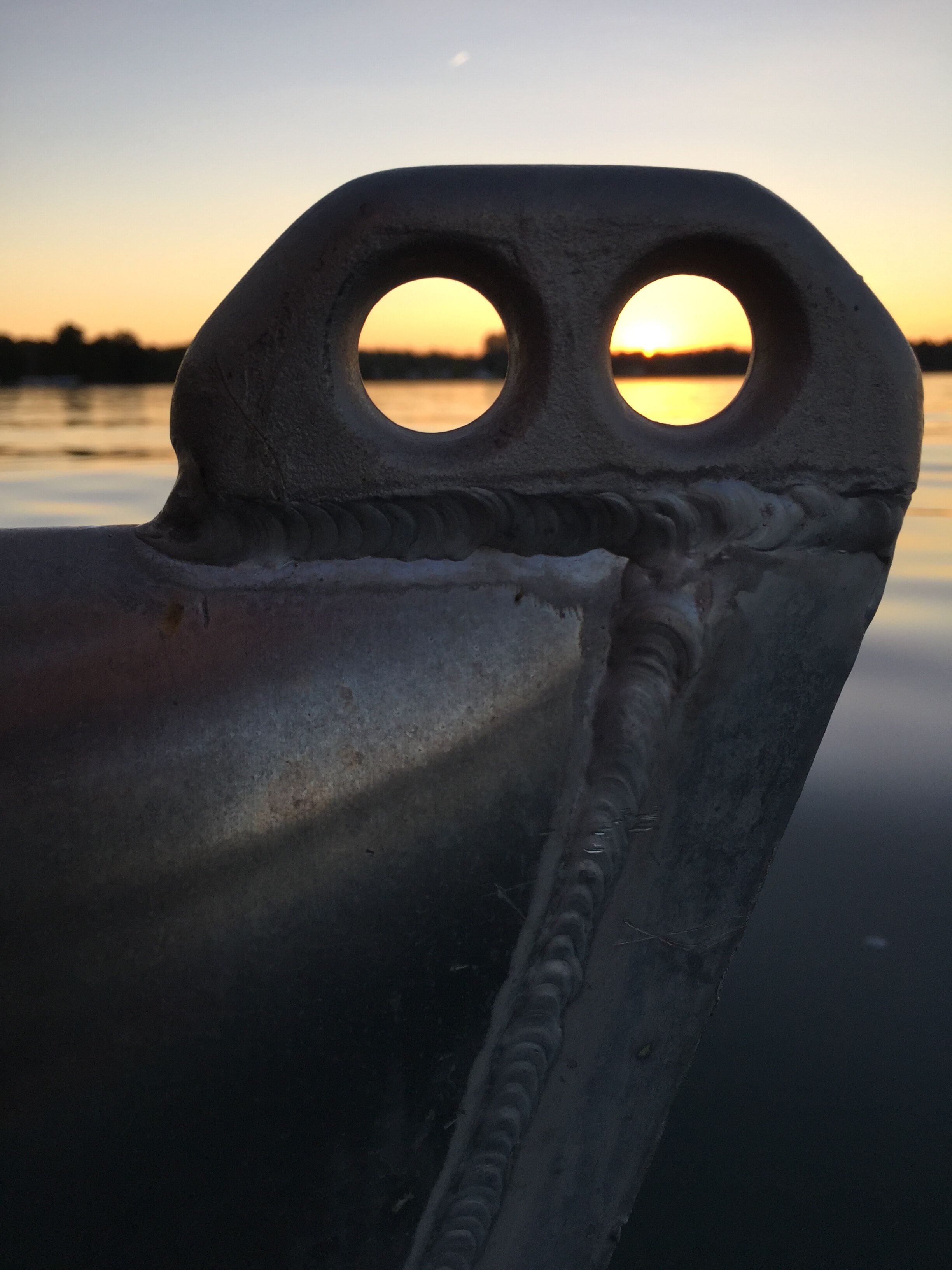 A metal structure with circular cutouts, likely part of a boat or dock, with a sunset in the background.